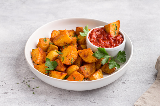 Traditional Spanish potato, patatas bravas with smoked paprika, spicy tomato sauce and parsley in a white bowl on a gray stone background, top view. Delicious homemade food