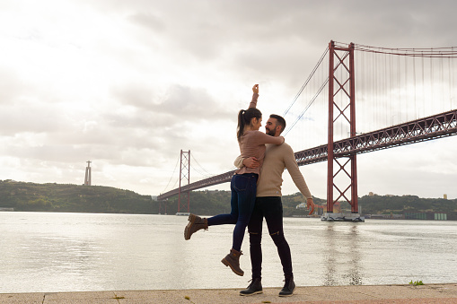 Man holding his partner while jumping, on the bank of the Tagus River next to the 25th of April bridge on an autumn day.