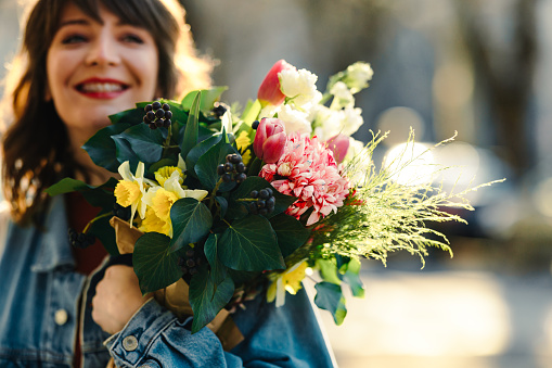 Cropped photo of a young cheerful woman walking in the city with a bouquet of flowers. She is smiling and looking away.