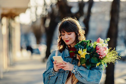 Young cheerful woman walking in the city with a bouquet of flowers. She is wearing denim jacket and a bag while texting.