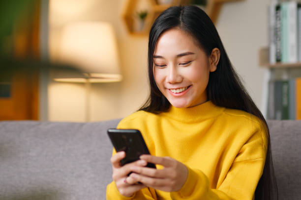 Young Asian beautiful woman sitting on sofa using smartphone, mobile for surfing internet, social media, online shopping, chatting with friends at home stock photo