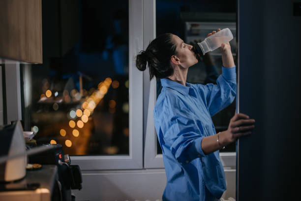 Woman holding the fridge door open while she drinks water from a bottle, at night in her kitchen at home.