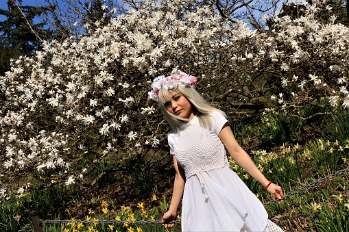 A Vietnamese model in a garden in front of a Magnolia tree. She is wearing a white headband and white layered dress.