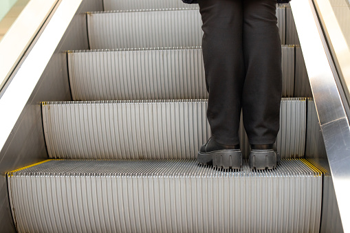 Legs in black pants on step of escalator in shopping mall. Metal staircase. Up escalator. Close-up.