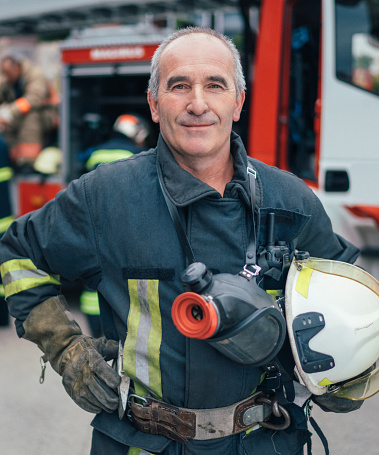 Portrait of a Caucasian fireman wearing his firefighter suit, standing next to a fire engine at the station. He is holding his firefighter helmet.