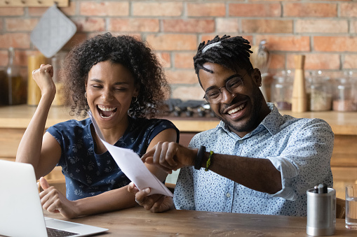 Happy excited young Black couple celebrating financial success, achieve, holding papers, reading document, making winner gesture, paying off mortgage loan, getting unexpected income, profit