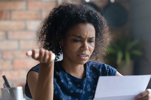 Frustrated millennial young Black woman reading paper letter with bad news. Mixed raced student girl receiving rejection notice from college, bank, reviewing document, feeling sad, upset