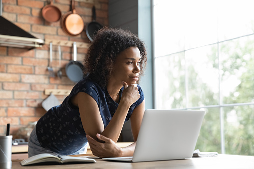 Thoughtful female African American remote employee standing by computer at home, leading on table, taking work break, looking at window in deep thought, thinking over project future vision,