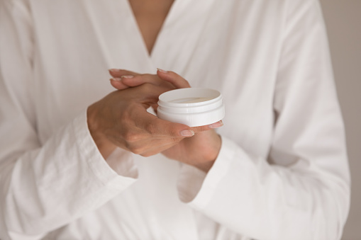 Close up young woman in white bathrobe holds jar, applying hand cream, caring for skin, use natural cosmetics product, gives skin pleasant fragrance, makes smoother, softer, younger. Skincare concept