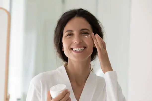 Head shot portrait happy Hispanic woman in white bathrobe smile looks at camera holds jar applying cream on cheek area. Anti-ageing and wrinkles cosmetics, skincare and beauty, morning routine concept