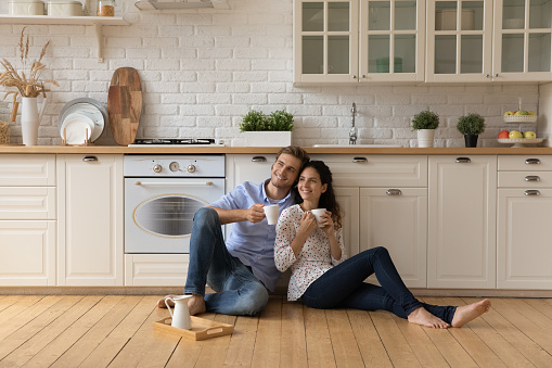 Homeowners family couple in love relax on wooden warm floor in kitchen holds cups drinking tea or morning coffee beverage enjoy daydream together at new own or rented house. Tenancy, bank loan concept
