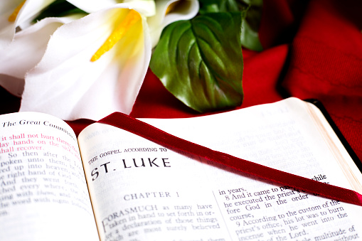 Bible Open to the beginning of the book of St Luke, New Testament.
Red satin bookmark is across the page.
Background is a White lily on red tablecloth.