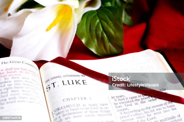 Bible Open To The Beginning Of The Book Of St Luke New Testament Red Satin Bookmark Is Across The Page Background Is A White Lily On Red Tablecloth Stock Photo - Download Image Now