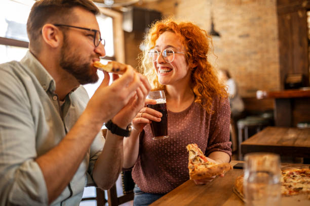 coppia sorridente felice che si diverte mentre mangia pizza e beve birra in un pub - pizzeria foto e immagini stock