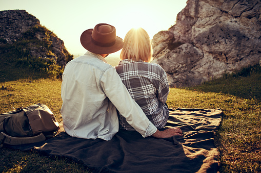 Rear view of young couple sitting on blanket in the mountains enjoying a beautiful sunset together