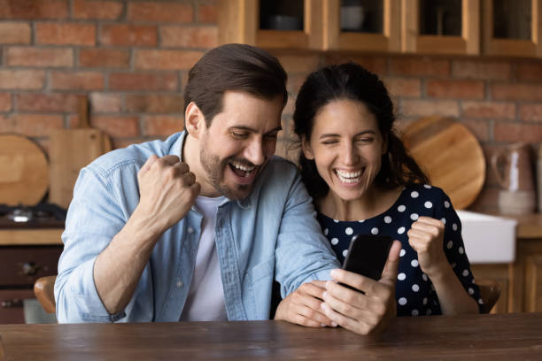istock Overjoyed young married couple hold smartphone yell yes at kitchen 1389746532