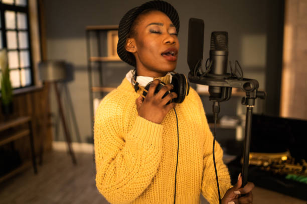 A behind the scenes look at a singer rehearsing in private, standing by a professional microphone and holding headphones around her neck, singing with her eyes closed