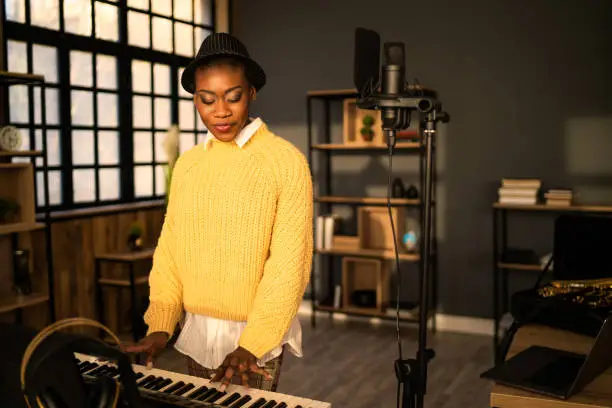 A female multiethnic entertainer is playing an electric keyboard in a dark music studio, wearing a bright yellow sweater and a porkpie hat