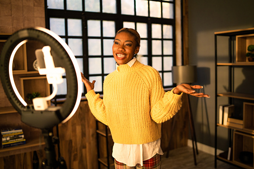 An Anglo-Caribbean woman is gesturing with her hands as she films her daily webcast on her cellphone attached to a ring light in a modern room at home