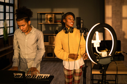Two entertainers singing and playing a keyboard for a musical video blog using a mobile attached to a selfie ring light set up in front of them while they enjoy their performance