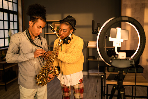 An Anglo-Caribbean woman is helping a saxophonist with his strap before rehearsals, there is a mobile phone set ready in an LED ring light ready to record for their vlog