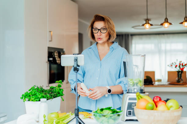 middle age women is blogging for her wellness lifestyle channel about healthy balanced living in the kitchen of her home. creating video content for social media with phone camera on a tripod. - television camera tripod media equipment videography imagens e fotografias de stock