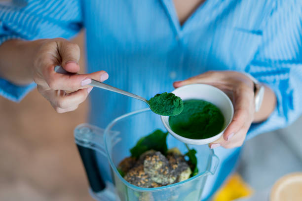 キッチンでスムージーを作る際にスピルリナグリーンパウダーを加えるクローズアップ女性。スーパーフードサプリメント。健康的なデトックスビーガンダイエット。健康的なダイエット、� - spirulina bacterium ground algae mineral ストックフォトと画像