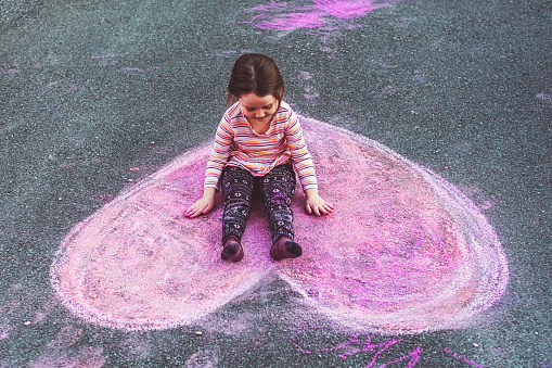 A five year old girl sits happily on a  chalk heart in her home's driveway.