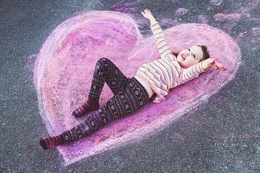 A five year old girl makes silly faces while lying on a  chalk heart in her home's driveway.