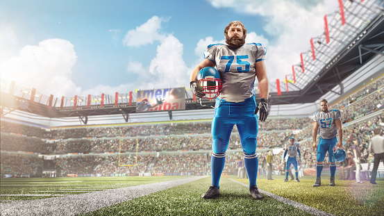 An American football player stands in stadium and holds a helmet in his hand. Stadium during the day. Sport. Man