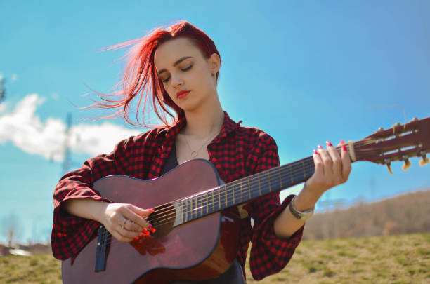 schönes teenager-mädchen mit gefärbten roten haaren, das an dem schönen sonnigen tag im freien braune akustikgitarre spielt - karohemd stock-fotos und bilder