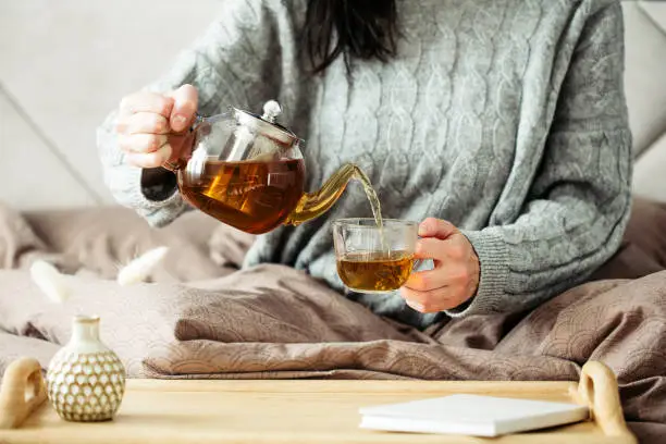 Photo of Woman dressed in knitted sweater pours hot tea from glass teapot into mug while sitting in bed. Morning breakfast in cozy home bedroom interior. Hygge, warm, autumn concept.