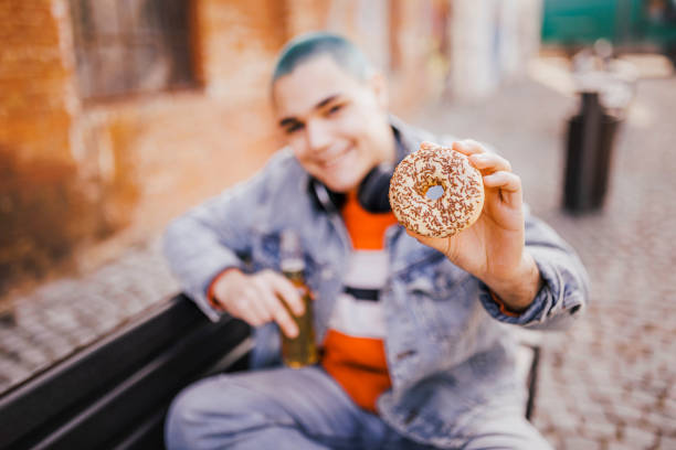 istock Smiling man drinking beer, eating doughnut and sitting on park bench 1389758040