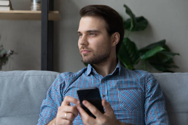 istock Man sits on sofa holds smartphone looking into distance 1389758502