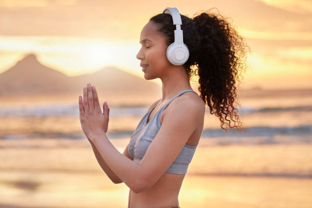 scatto di una donna che indossa le cuffie mentre pratica yoga sulla spiaggia - real estate immagine foto e immagini stock