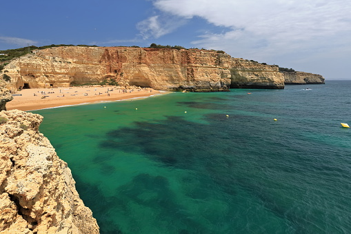Praia de Benagil Beach seen from the cliff framing its west side backed by the vertical walls pierced by caves and holes of the yellowish to pinky geological Lagos-Portimao Formation. Algarve-Portugal