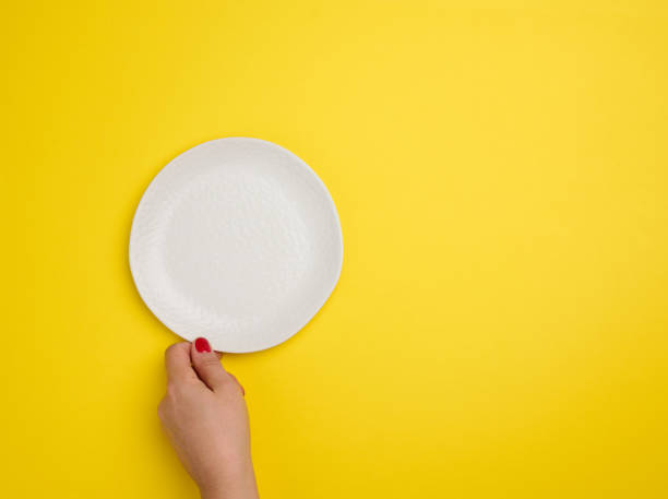 istock female hand holding a white empty round plate on a yellow background, top view 1389764404