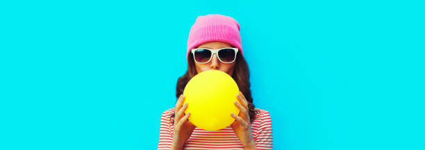 Fashionable portrait of stylish cool young woman inflating chewing gum wearing pink hat on blue background stock photo