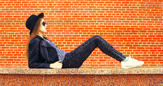 Stylish young woman sitting in the city wearing black rock style over brick wall background