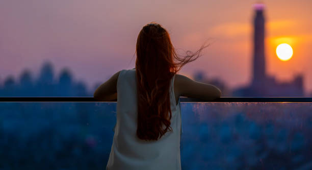 istock Woman looking and enjoying the sunset view from balcony with the sun setting behind skyscraper in busy urban downtown with loneliness for solitude, loneliness and dreaming of freedom lifestyle 1389774793