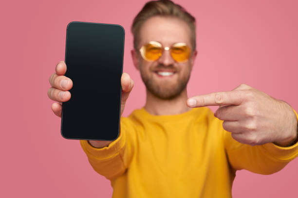 Cheerful male hipster in bright outfit showing mobile phone and pointing at black display on pink background in studio