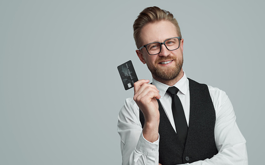 Cheerful respectable male entrepreneur showing black credit card for online payment and looking at camera on gray background in studio