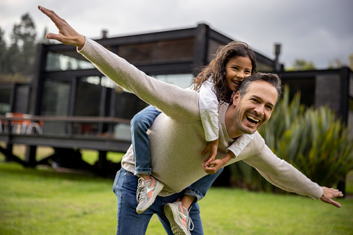 Happy Latin American father having fun playing airplane outdoors with his daughter and smiling - lifestyle concepts