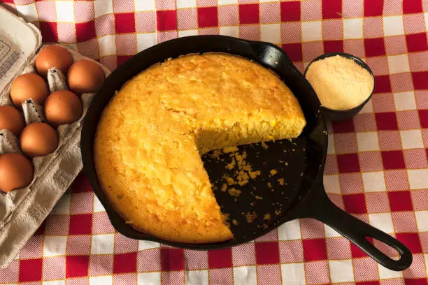 Homemade cornbread in a cast iron skilet, with eggs and a container of corn flour arranged on the side.