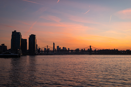 A colorful sunset along the East River in New York City with the Williamsburg Bridge and skyscrapers in the distance