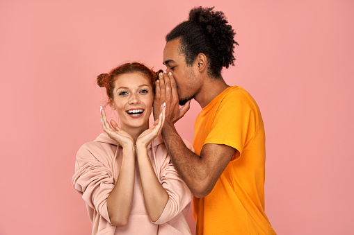 Cheerful black guy whispers secret to ginger caucasian girlfriend shares gossip or secret have fun, who has cheerful expression, gossip together, stands against pink background. Diverse couple indoor.