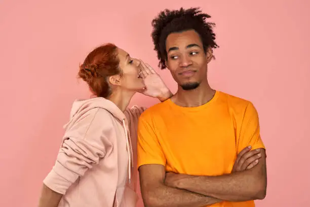 Photo of Ginger female whispers secret to Afro American boyfriend who has cheerful expression gossip together