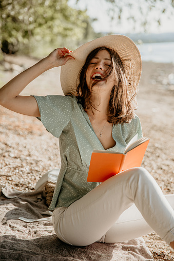 Fashionable glowing, casual elegant dressed female holding a cute hat on her head, smiling, enjoying a sunny windy day by the water/river. Vacation and lifestyle concept. She is reading and enjoying the sun