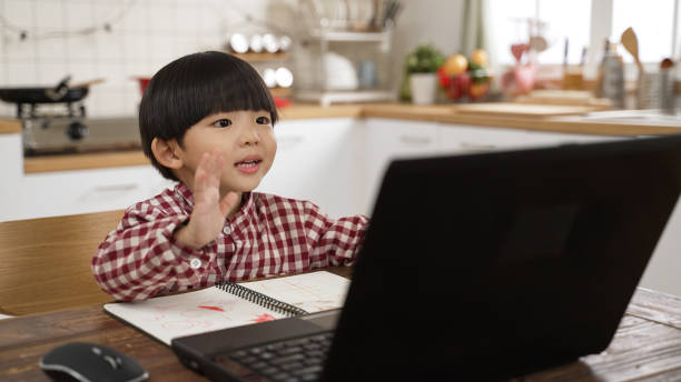 cute Asian schoolboy waving hi to laptop screen while taking online lesson in the dining room at home. learning from home concept cute Asian schoolboy waving hi to laptop screen while taking online lesson in the dining room at home. learning from home concept Online Childcare Training stock pictures, royalty-free photos & images