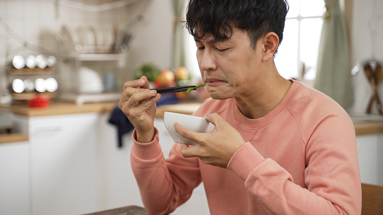 asian Japanese man grimacing face while trying a piece of vegetable in dining room at home. he finds the food too salty and puts down chopsticks